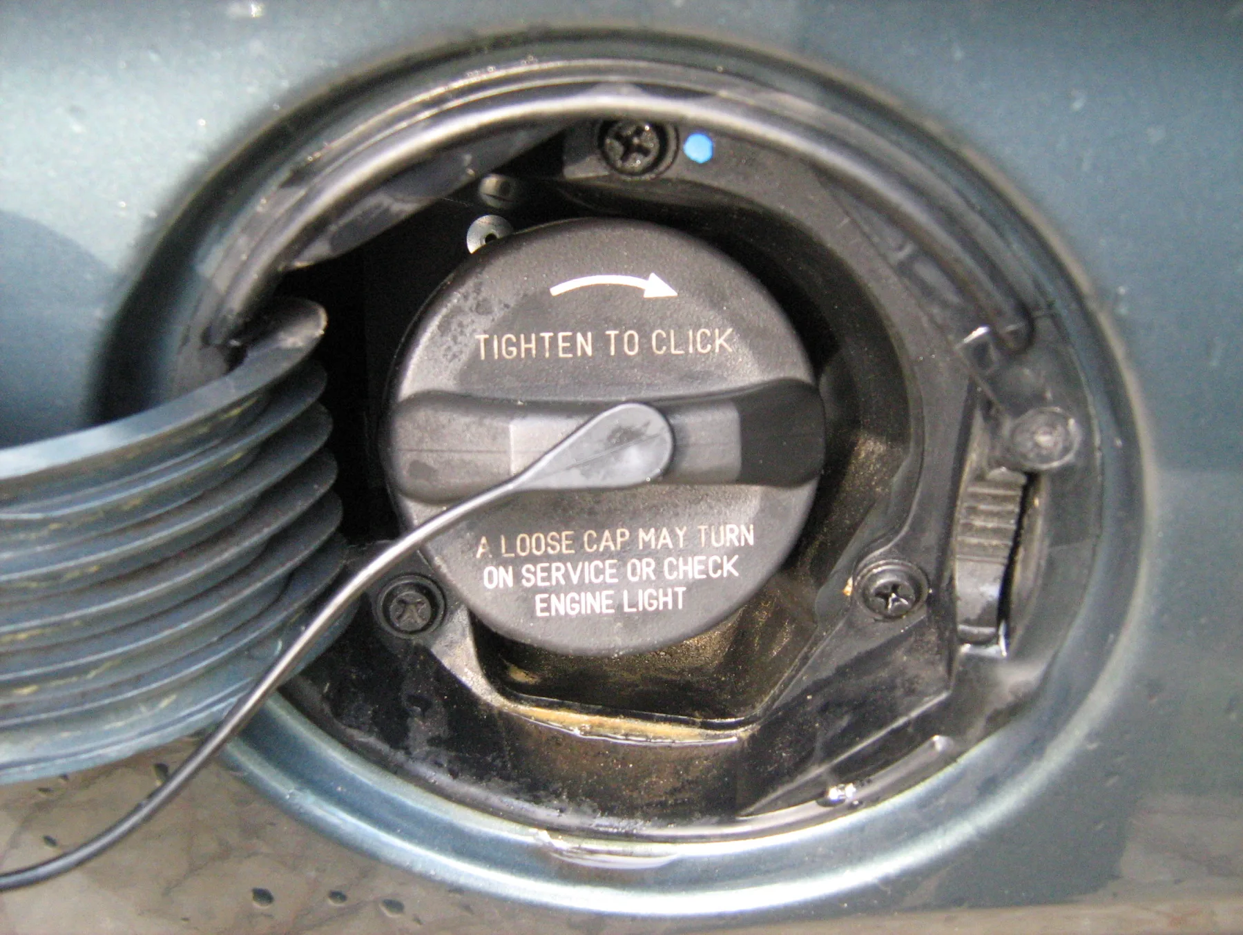 Black fuel cap seated in the filler neck behind an open fuel door, a thin wire tether running across it, molded text reading TIGHTEN TO CLICK with a curved arrow and A LOOSE CAP MAY TURN ON SERVICE OR CHECK ENGINE LIGHT
