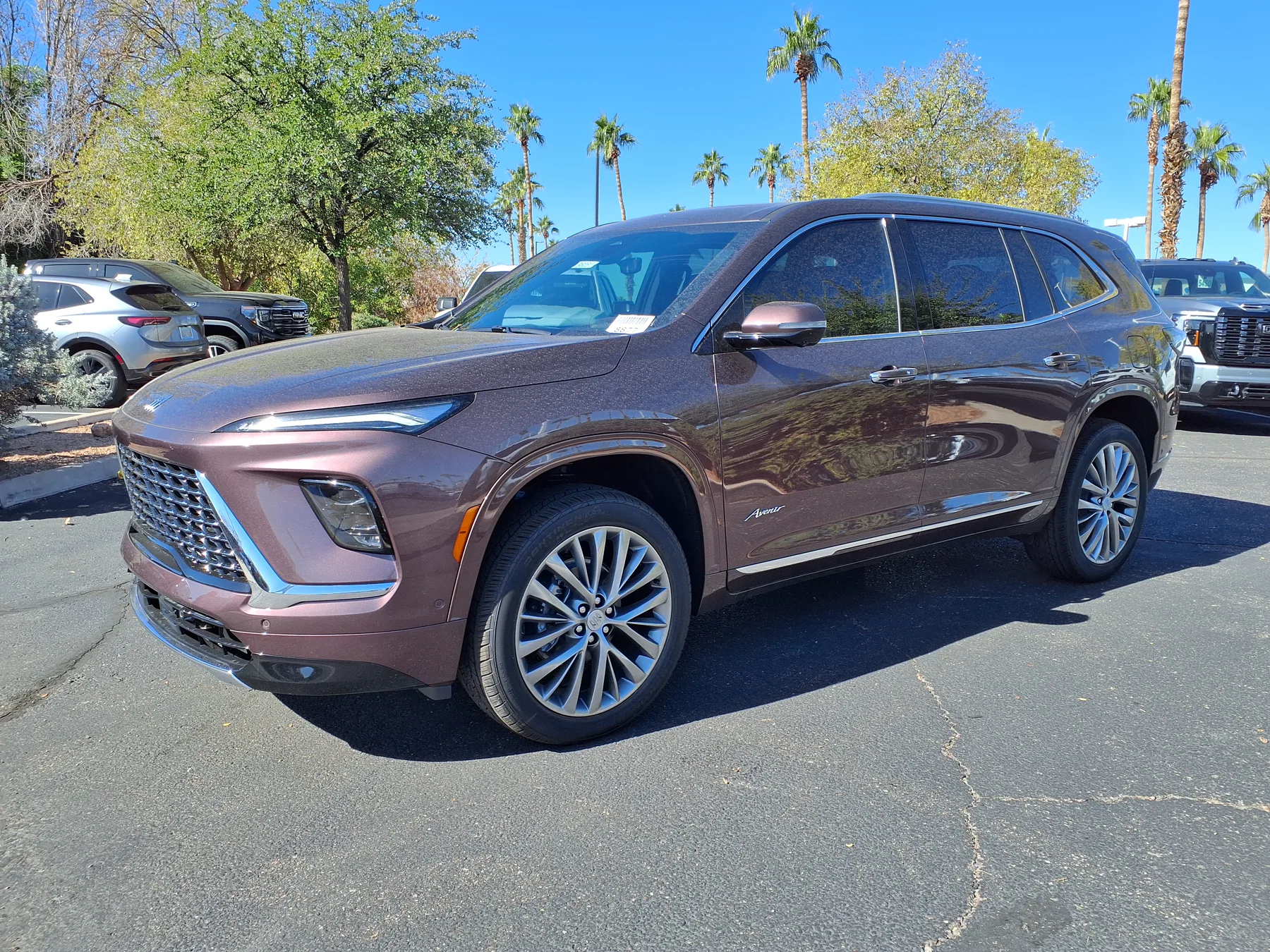 Front three-quarter of a 2026 Buick Enclave Avenir on a dealer lot, with check-shaped daytime lamps and a dark mesh grille