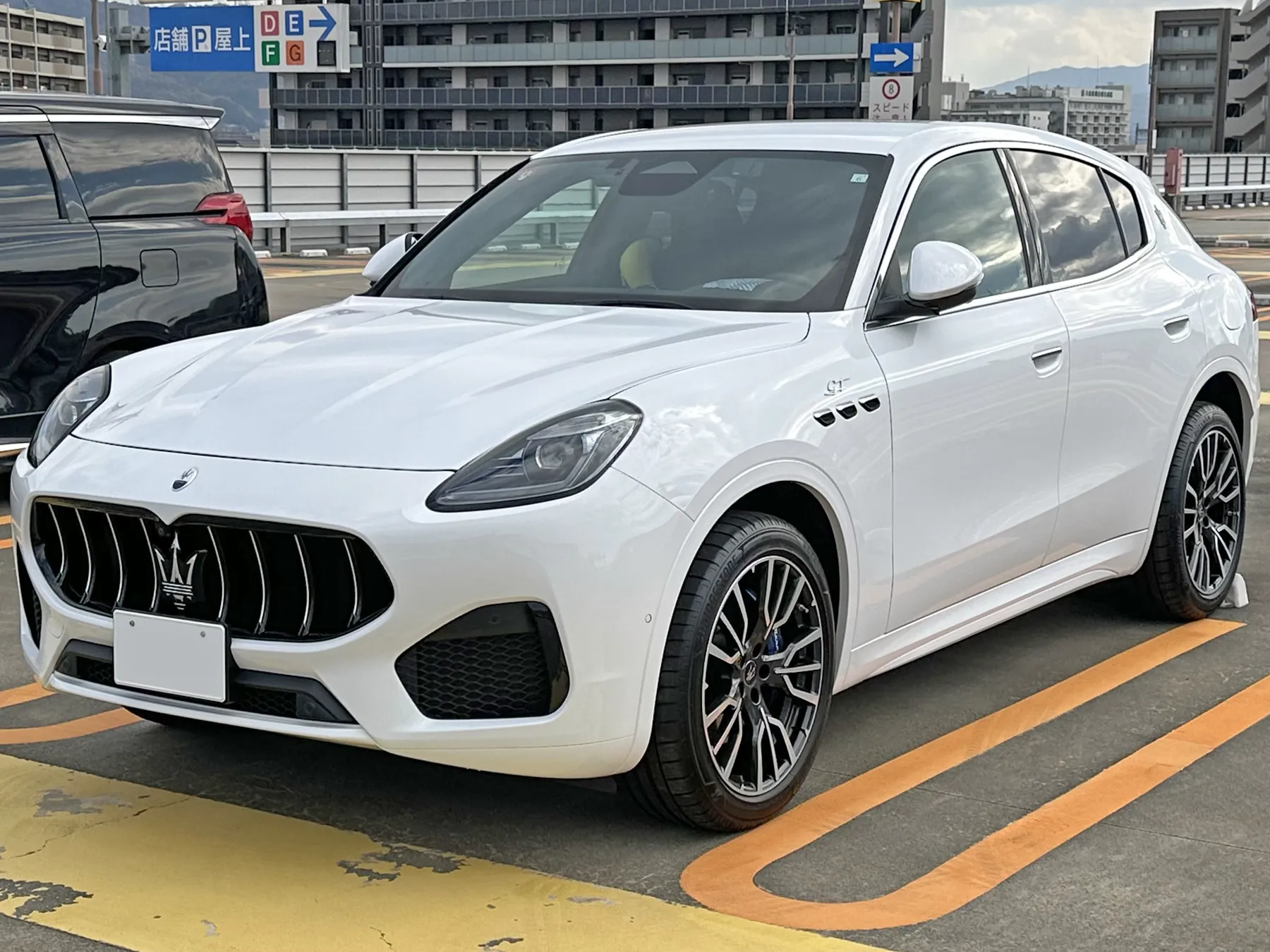 Front three-quarter of a white Maserati Grecale GT from the driver's left, outdoors on a rooftop parking deck with orange and yellow lines, a dark minivan at the left, and buildings and a blue Japanese parking sign behind. The grille has vertical dark and chrome bars with a chrome trident in the center, a blank white plate, a small round Maserati badge on the nose, slim headlamps, three small black vents on the front fender with small GT script above them, and dark multi-spoke wheels with blue calipers