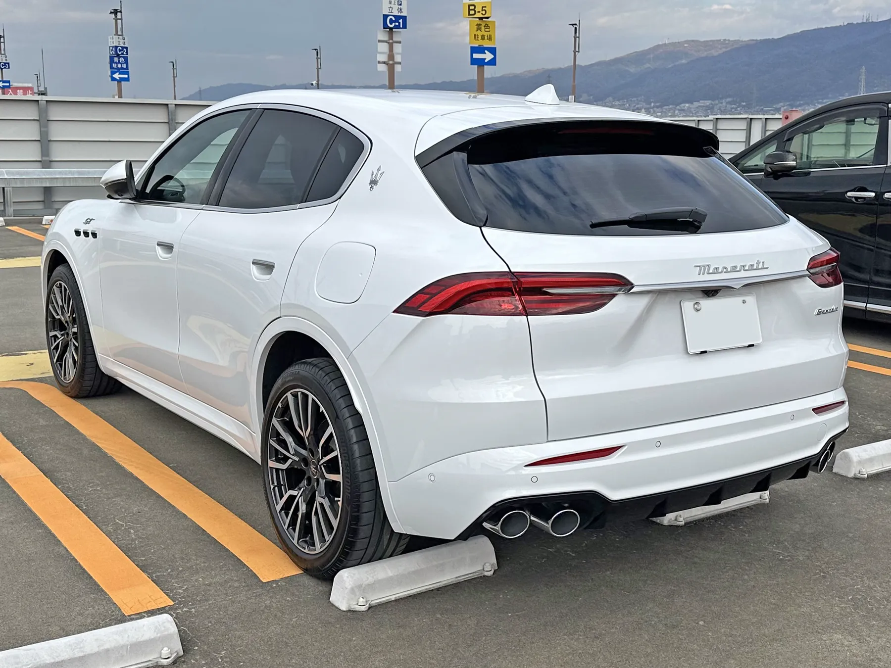 Rear three-quarter of the same white Maserati Grecale GT from the left, outdoors on the parking deck with a mountain range behind. Slim red tail lamps wrap from the tailgate into the body sides, Maserati is written in chrome script on the tailgate, the plate is blank white, a small trident badge sits on the C-pillar, a small script badge is at the right of the tailgate, four exhaust tips are in the bumper, and three fender vents show at the far left