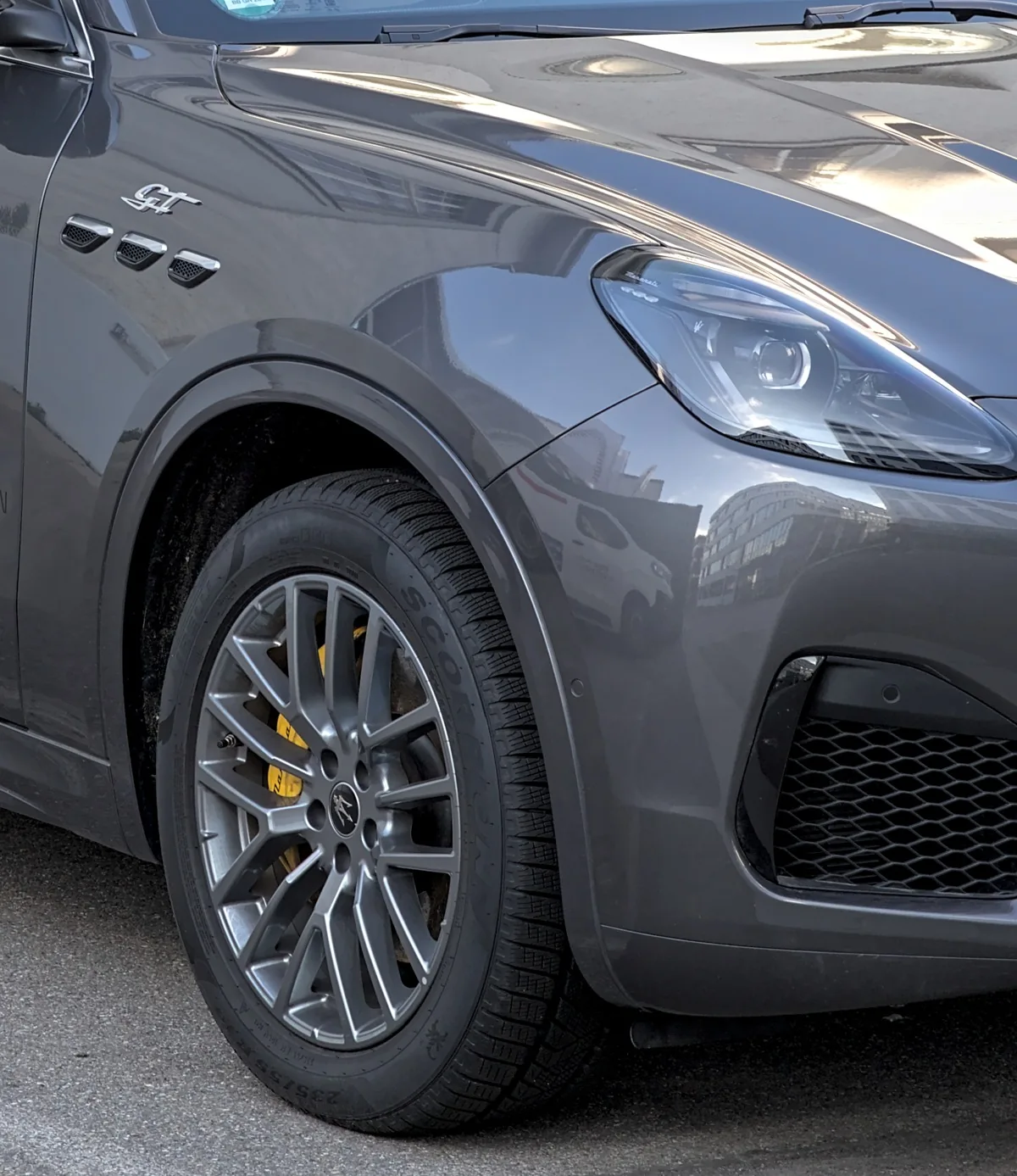 Front fender of a dark gray Maserati Grecale GT from the driver's left front. Three small chrome-edged vents with dark mesh sit side by side in a row on the fender, with a chrome GT script badge above them. The front wheel below is a silver multi-spoke wheel with a trident center cap and a yellow brake caliper behind the spokes, on a winter tire. On the right are the slim headlamp and part of a lower bumper intake with black hexagon mesh. Gray asphalt is below.