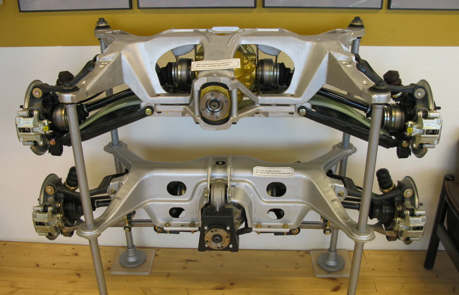 Two Volvo rear suspensions on display, the upper one with a light green transverse spring across the links and the lower one a multilink assembly without its coil springs