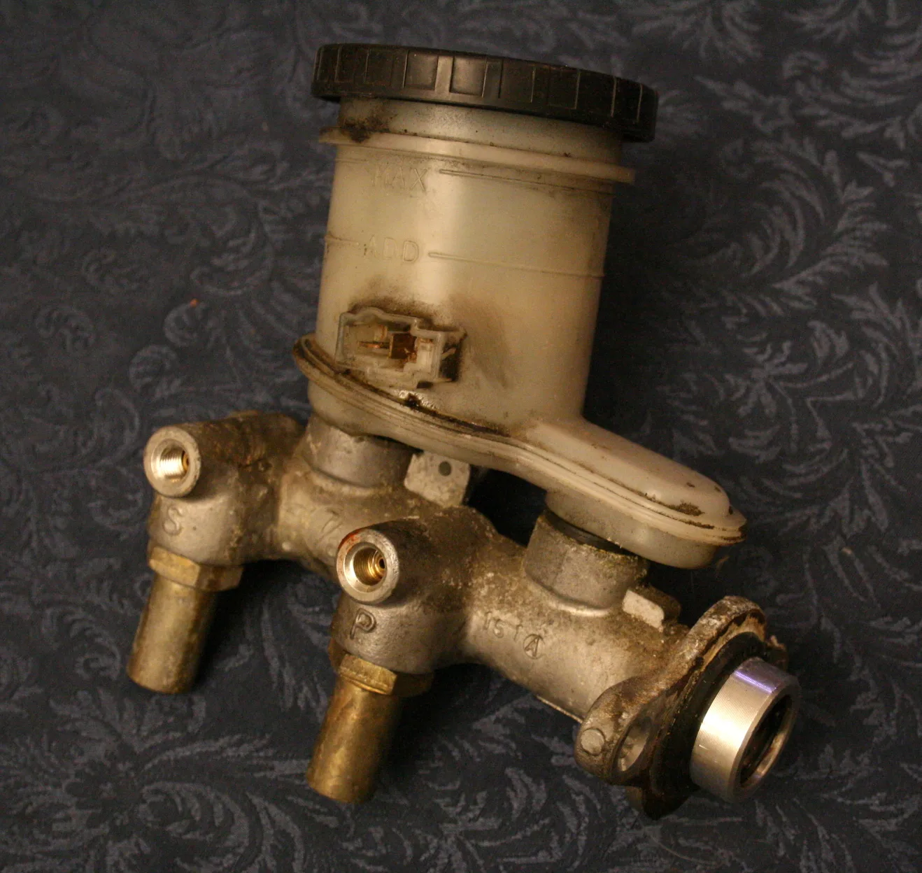 Removed metal brake master cylinder with a fluid reservoir on top and threaded connections in the body