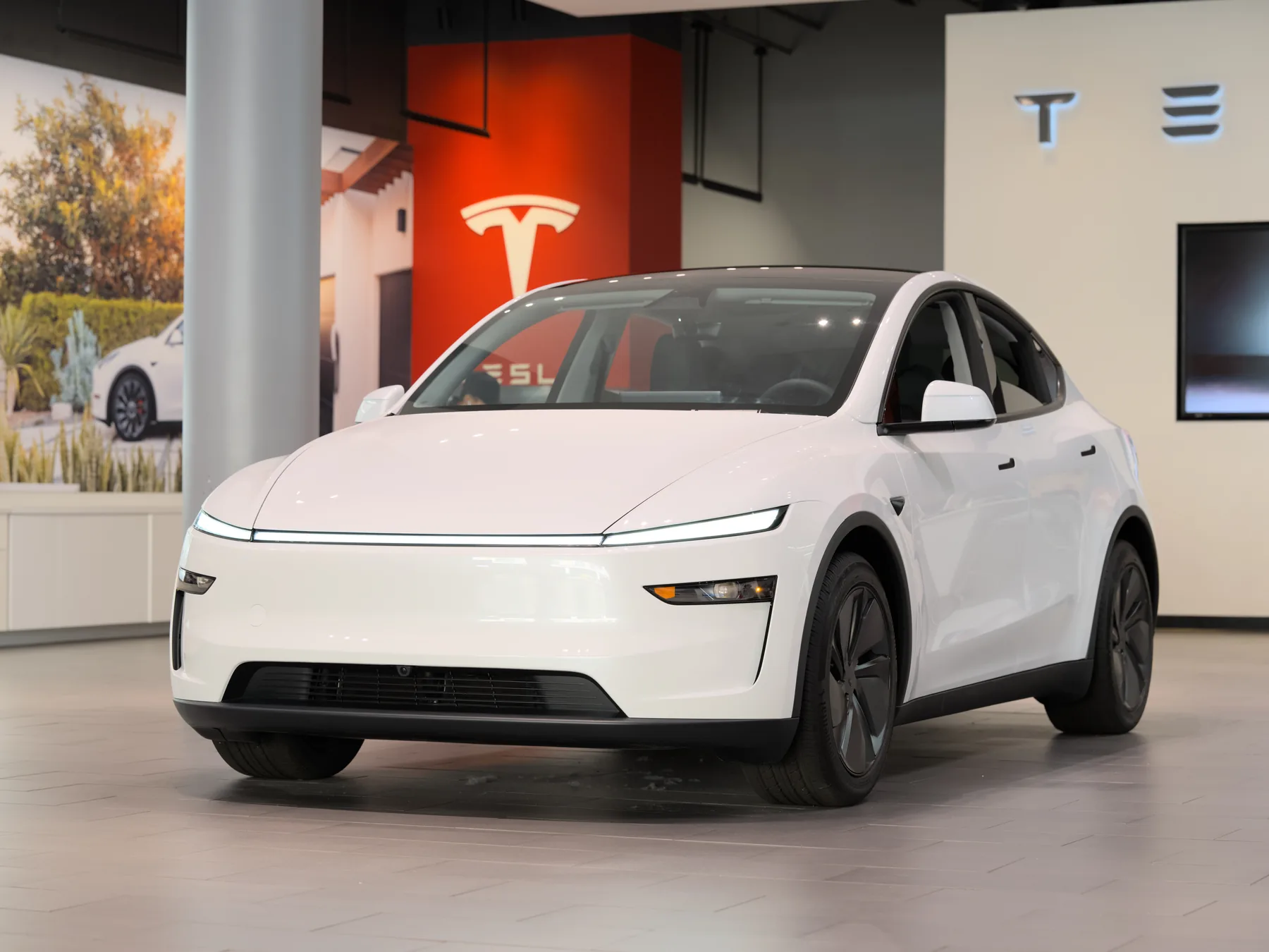 Front three-quarter of a white Tesla Model Y indoors in a showroom, with a lit thin light bar across the nose, slim headlamps low at the outer corners, a black lower intake, and a red wall with a Tesla T and TESLA lettering behind it