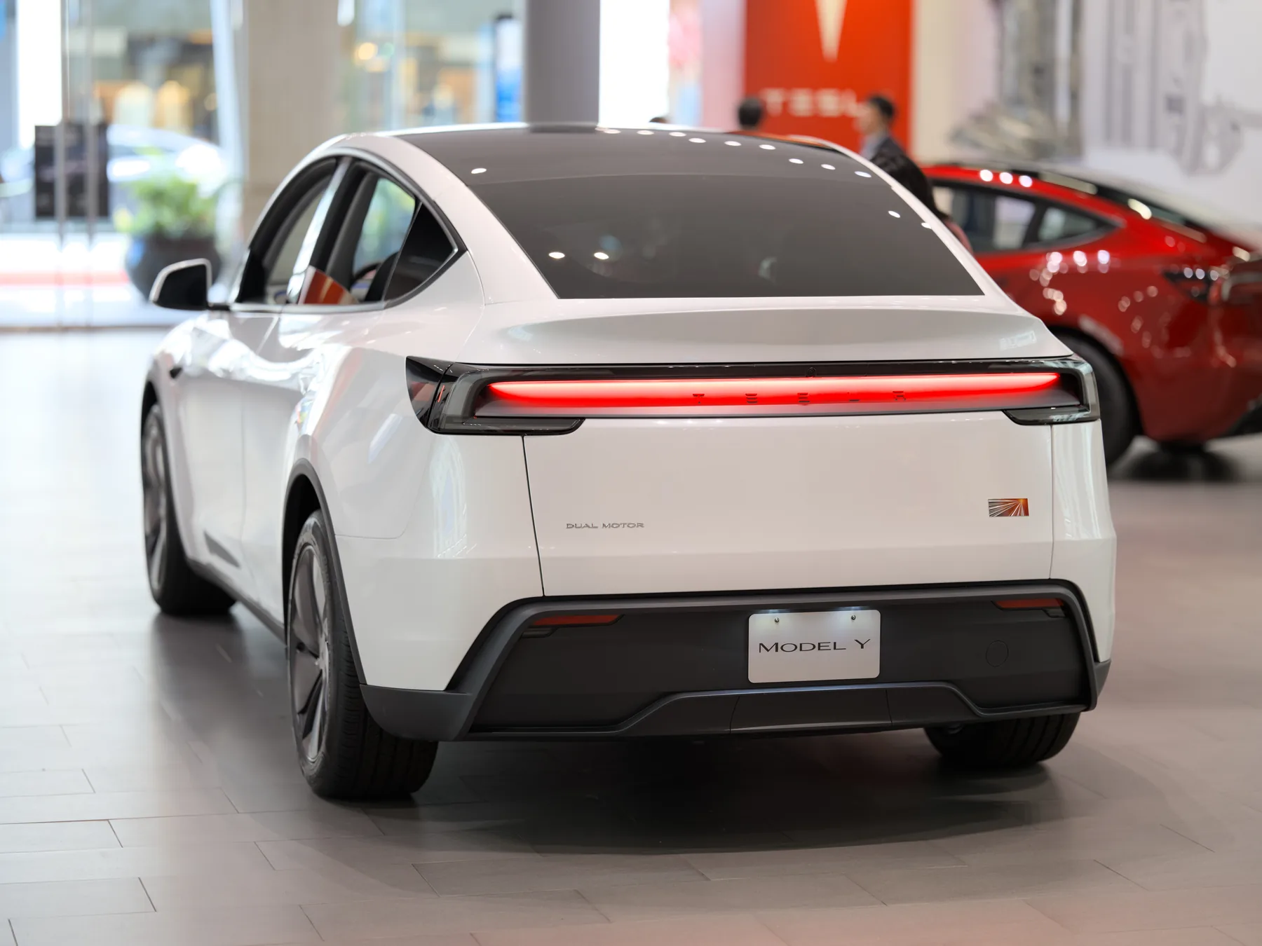 Rear three-quarter of a white Tesla Model Y indoors in the same showroom, with a lit red light bar across the tailgate, a DUAL MOTOR badge on the gate, a MODEL Y display plate, and a red Tesla in the background