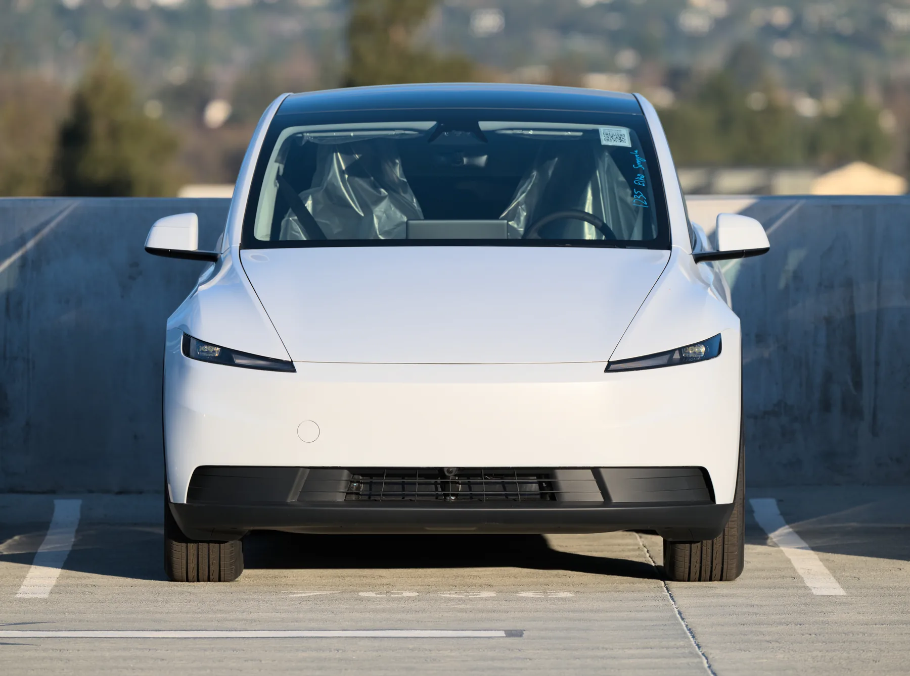 Head-on view of a white Tesla Model Y Standard outdoors on a parking deck against a concrete wall, with slim headlamps at each corner, a smooth body-colored nose and no light bar, a black lower intake, and seats wrapped in plastic