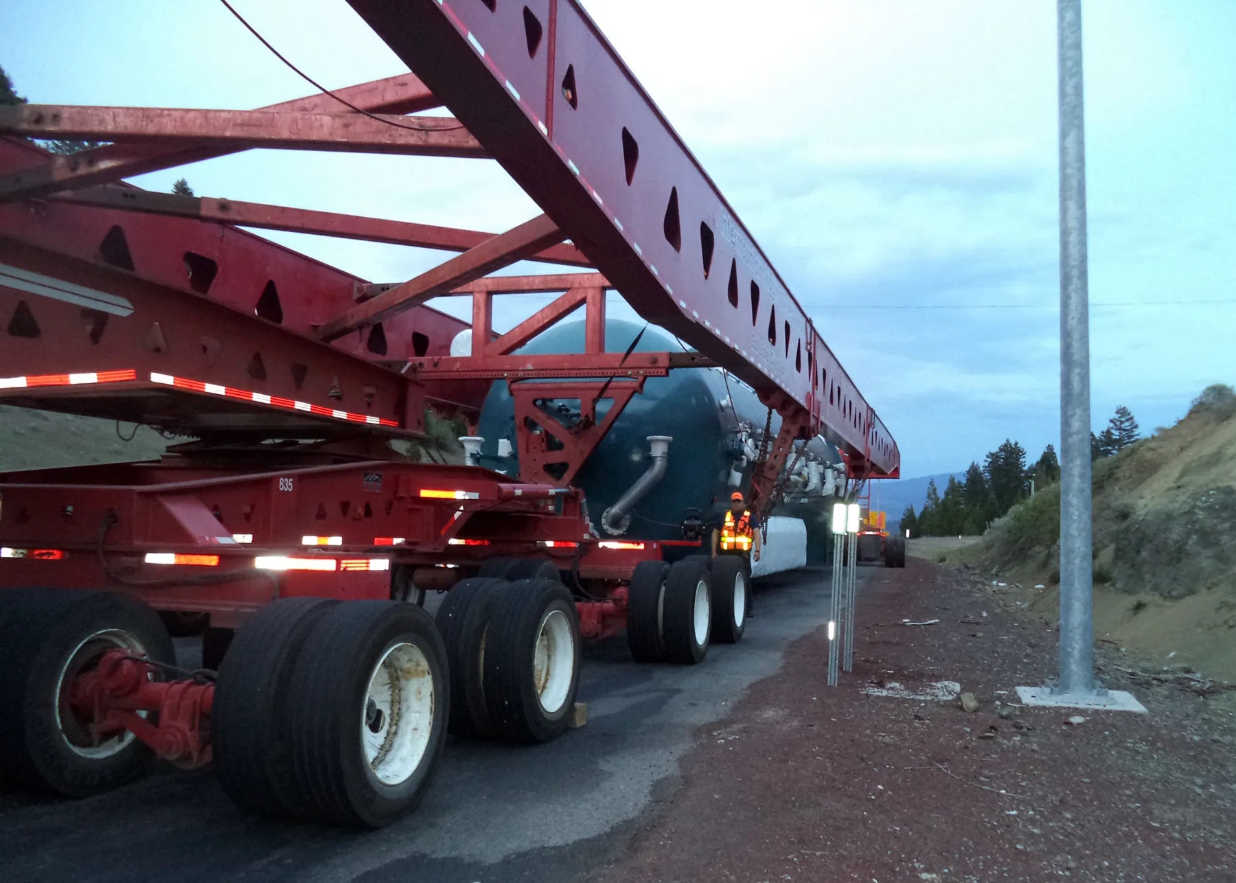 A view from the rear left, at dusk, of a red heavy-haul trailer. It has a long open girder frame with triangular cutouts overhead and rows of dual tires, and it carries a large dark green cylindrical vessel. A worker in an orange high-visibility vest stands beside the trailer. On the right are a gravel shoulder, a tall metal pole, white delineator posts, and a pine-covered hillside.