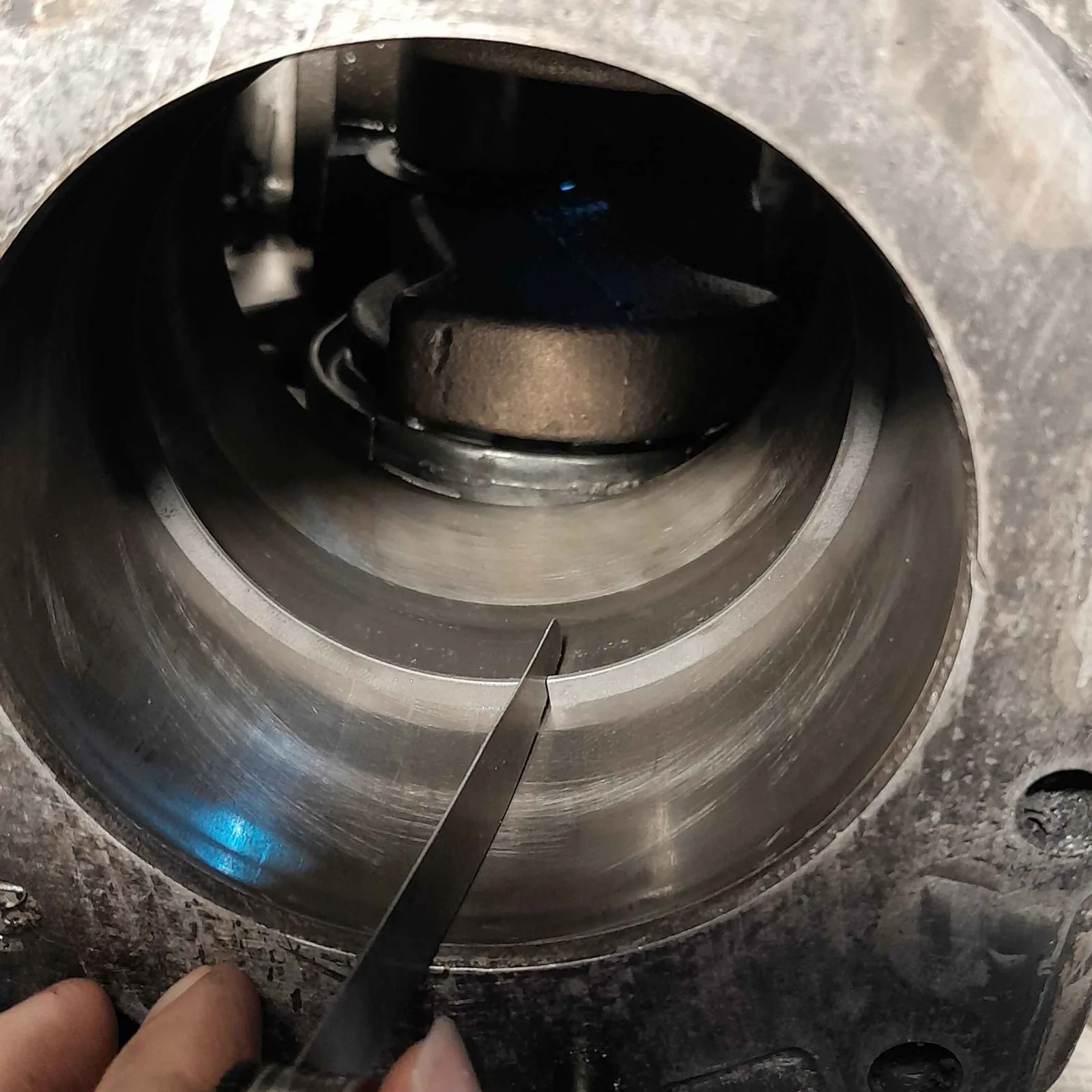 Looking down into a bare cylinder bore. A ring sits squared in the bore partway down, its ends parted. A single steel feeler-gauge blade, held by fingers at the bottom of the frame, is inserted in the end gap. The crankshaft and the connecting-rod end show at the bottom of the bore.