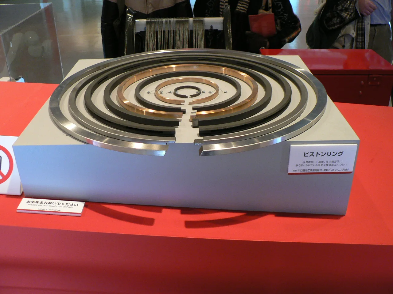 A trade-show display: a white block on a red table holds piston rings of many sizes, nested concentrically and laid flat. The outer rings are silver or dark gray, and the inner ones are copper colored. Every ring's open end gap faces the front. A small card at the right is labeled in Japanese, ピストンリング. Visitors' legs show behind the table.