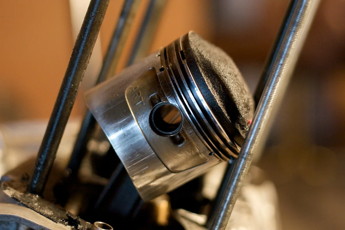 One piston hangs tilted on its connecting rod between engine studs, against a blurred orange background. The crown is black with carbon. Three rings sit in the grooves just below the crown. The wrist-pin bore is in the skirt, and a stamped mark is on the pin boss.