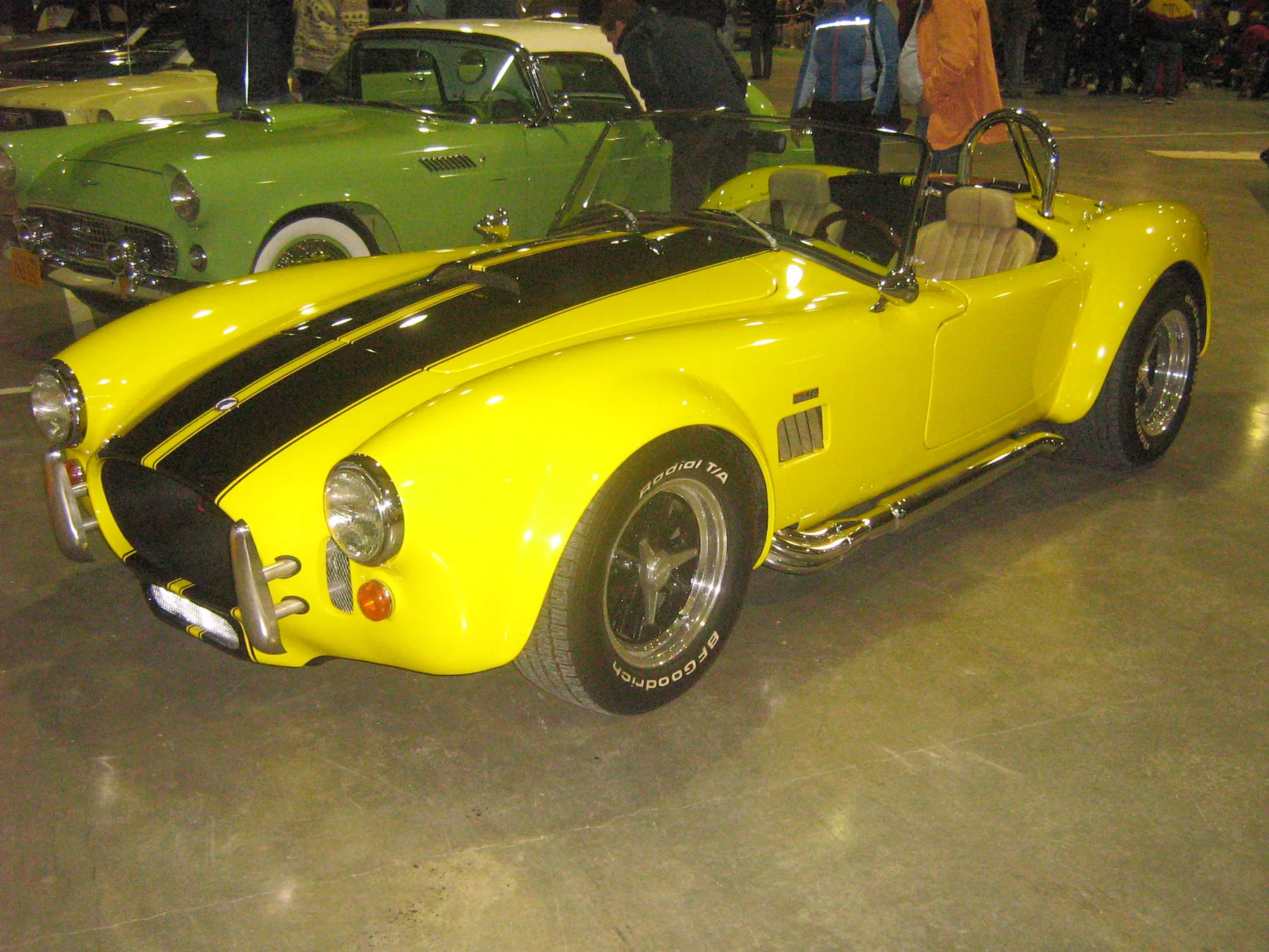 Yellow open two-seat sports car with black center stripes, a chrome roll hoop, and side pipes, indoors at a show with a green 1950s car behind