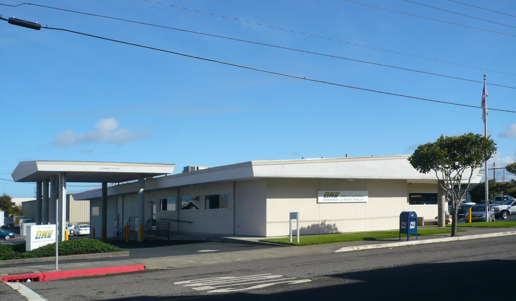 Long single-story beige DMV office under a blue sky, with a flat white roof edge and a covered drive-up canopy on the left, a wall sign reading DMV and State of California Department of Motor Vehicles, a DMV monument sign, a blue USPS mailbox, one flagpole with a U.S. flag, a tree, cars parked at the right, and power lines across the sky