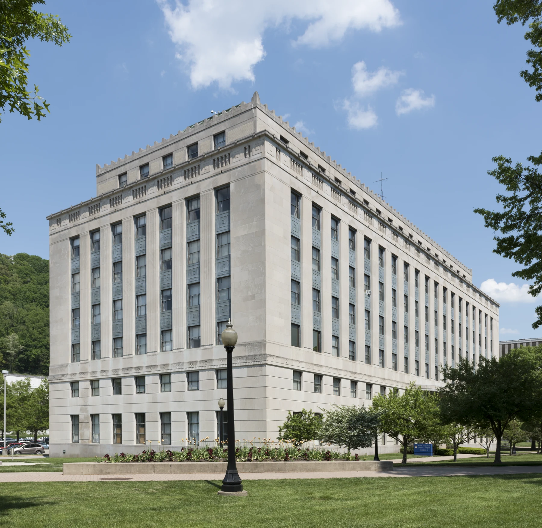 Tall many-story pale limestone office building with vertical bands of windows, seen at an angle across a lawn with a black lamppost and a flower planter in front, trees on both sides, and a wooded hill behind at the left under a blue sky