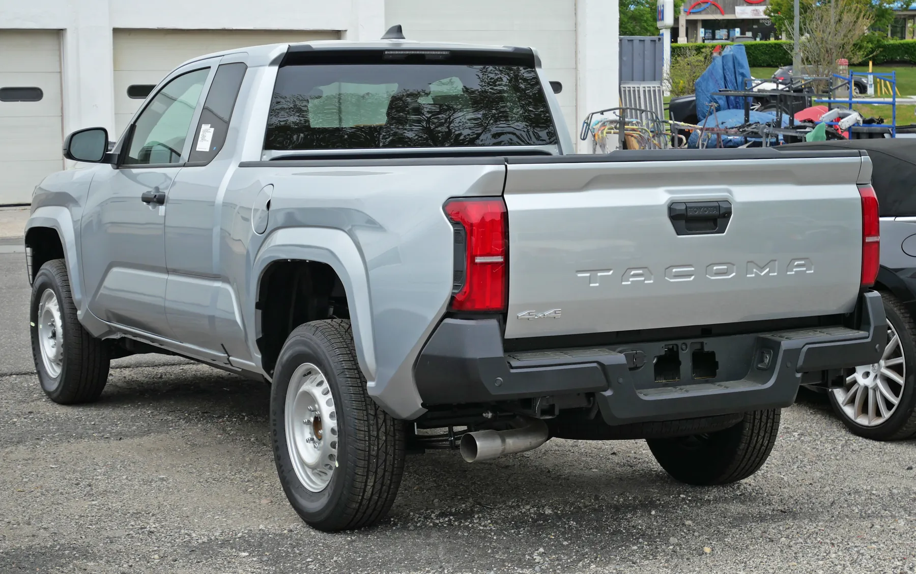 Rear three-quarter view from the left rear corner of a light gray Toyota Tacoma SR XtraCab, with a white garage building, roll-up doors, and trees behind. The gray tailgate has TACOMA pressed across the center under a black handle and a 4x4 badge at the lower left of the gate. The top edge of the tailgate is dark. The center brake lamp sits at the top of the cab. A vertical red taillamp is on the left bedside corner. The rear bumper is black, silver steel wheels are visible, and an exhaust tip shows under the bumper. The left bedside has no grade badge.