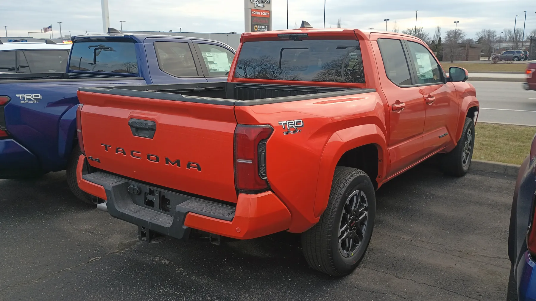 Rear three-quarter view from the right rear corner of an orange Toyota Tacoma TRD Sport Double Cab on a dealer lot. A blue Tacoma TRD Sport is parked beside it on the left, with light poles and a store sign behind. The orange tailgate has TACOMA pressed across the center under a black handle and a small 4x4 badge at the lower left of the gate. The top edge of the tailgate is black. The center brake lamp sits at the top of the cab above the rear window. A vertical red taillamp is on the right bedside corner beside the gate. The rear bumper is dark gray with orange end caps. A TRD SPORT badge is on the right bedside, and a dark alloy wheel is visible on the right side.