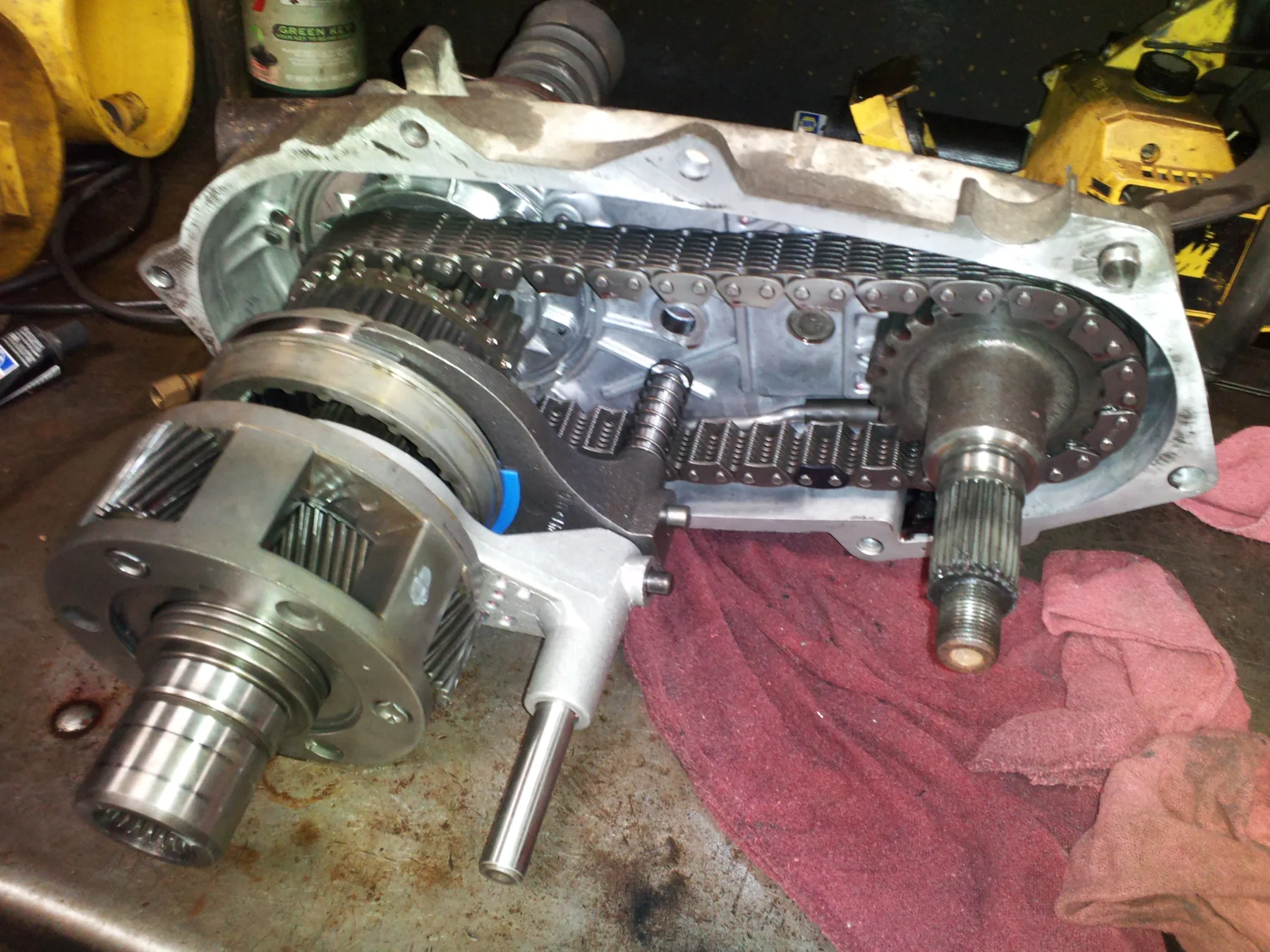 Opened chain-drive transfer case on a workbench beside a red shop rag, case half off, a chain between two sprockets, a planetary gearset and shaft at the left front, a shift fork and rail in the middle, and a splined output shaft at the right