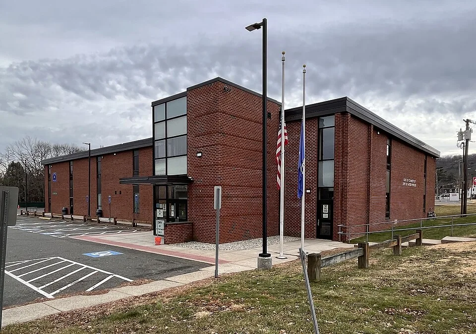 Two-story red brick building with dark flat-roof trim and a tall glass stair tower at the entrance, two flagpoles with a U.S. flag at half-staff and a blue flag, a tall black lamppost, a parking lot with white stripes and a blue accessible space at the left, a wooden rail fence and brown winter grass in front, an overcast gray sky, and bare trees